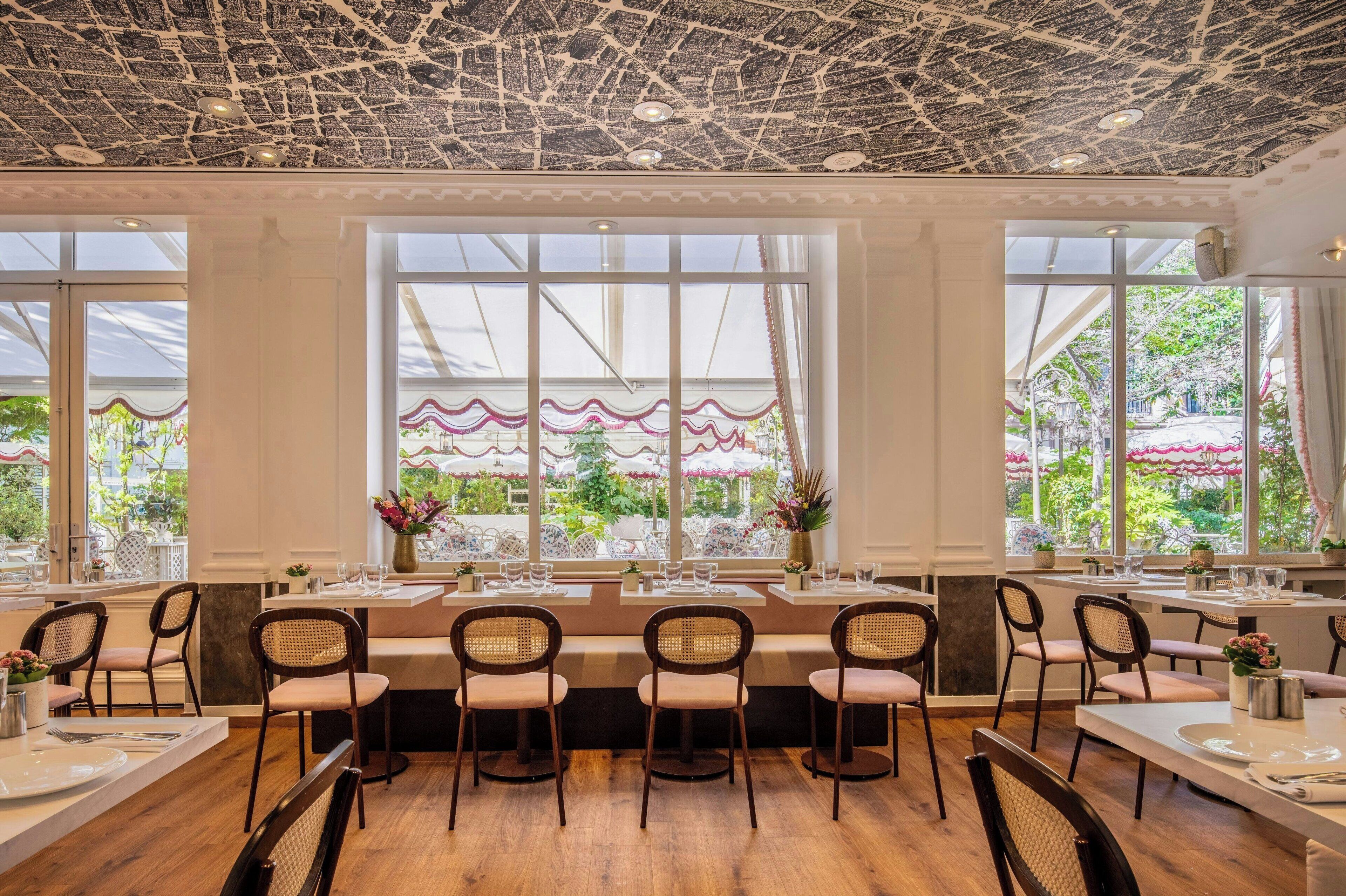 A restaurant with a high ceiling, large windows, and a view of a garden. Tables are set with chairs and centerpieces.