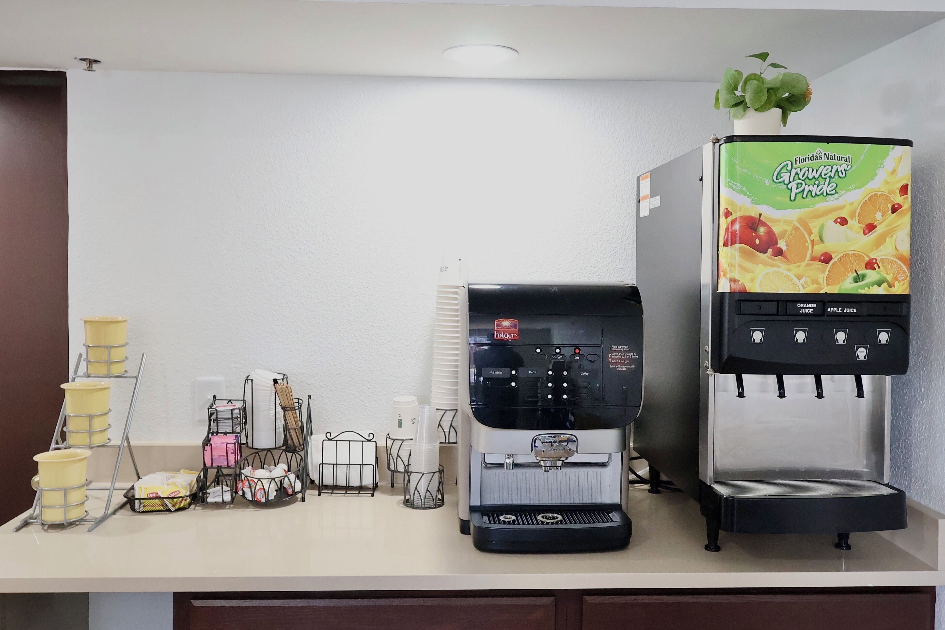 Un coin café équipé d’une machine à café et d’un distributeur de jus, de tasses et de serviettes, ainsi que d’une petite plante fixée au mur.