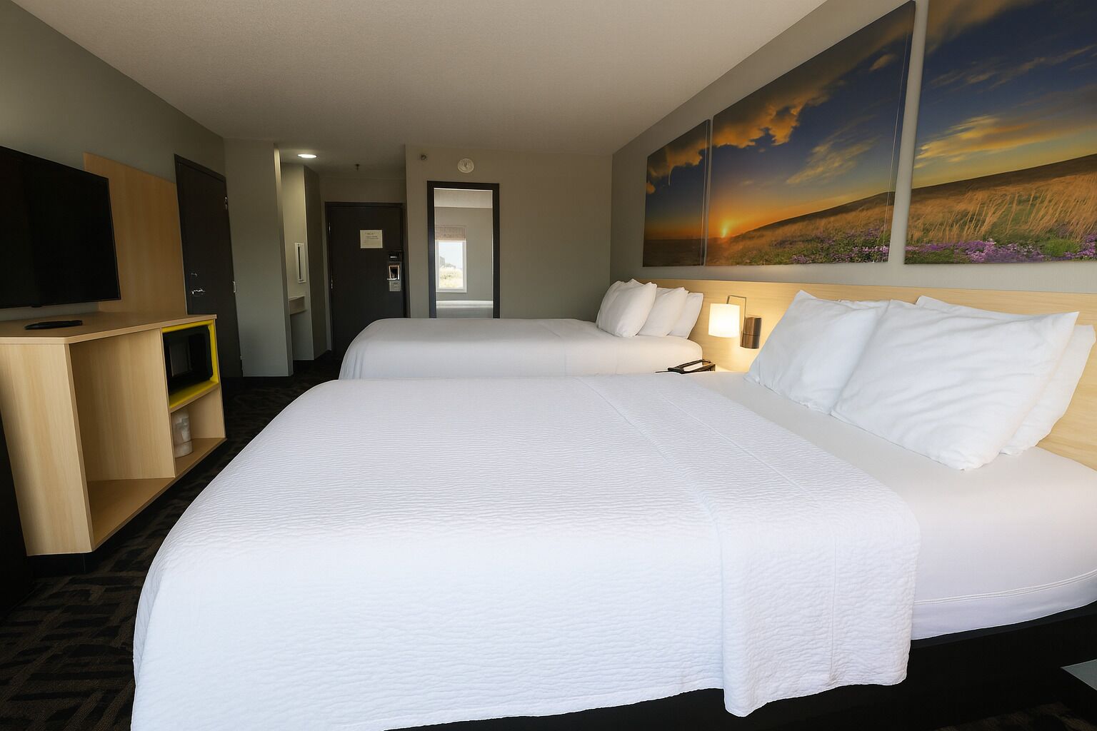 Quarto de hotel com duas camas, televisão, escrivaninha e uma pintura de pôr do sol.