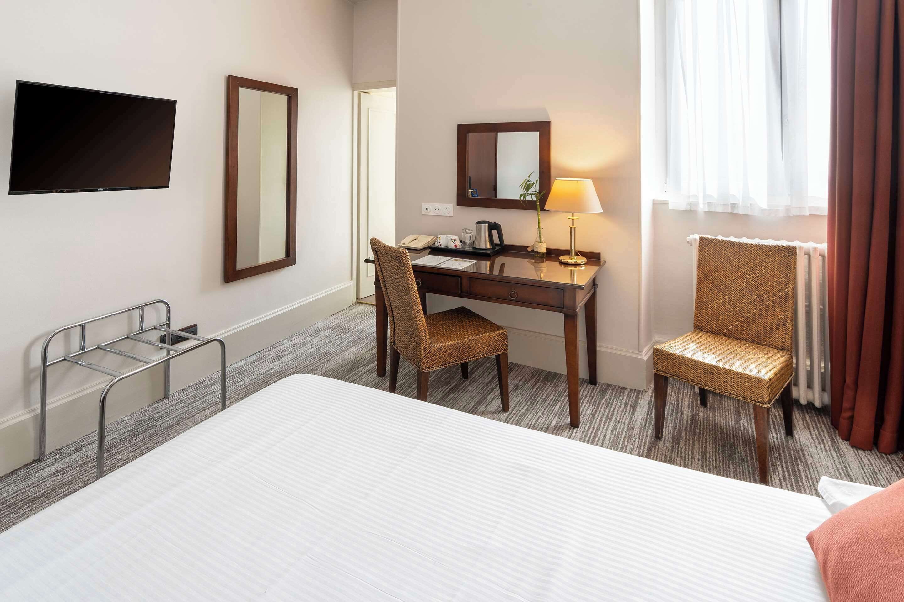 A hotel room with a bed, a desk with a chair, a television, a mirror, and a lamp.