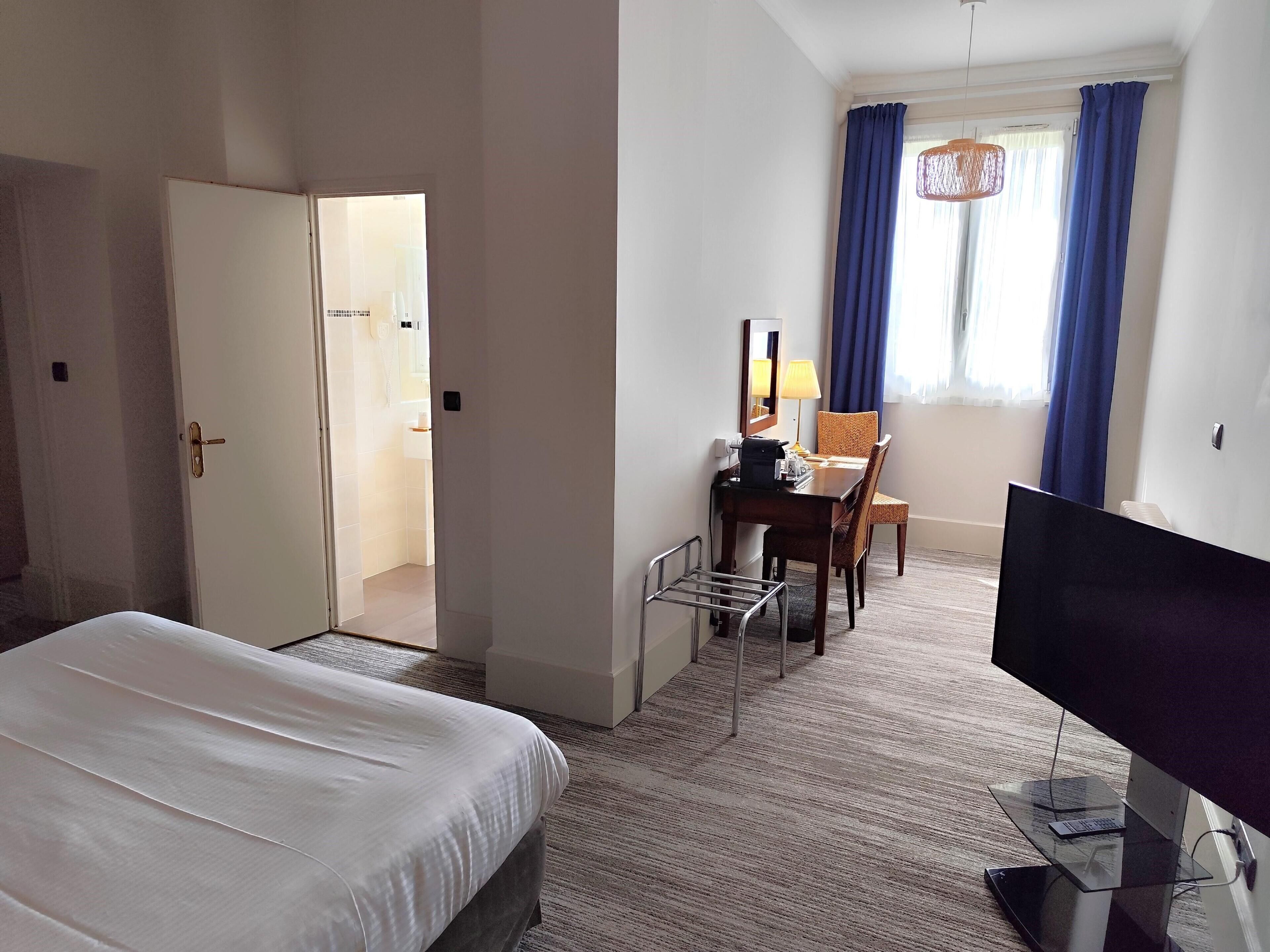A hotel room with a bed, a desk with a chair, a television, and a window with curtains.