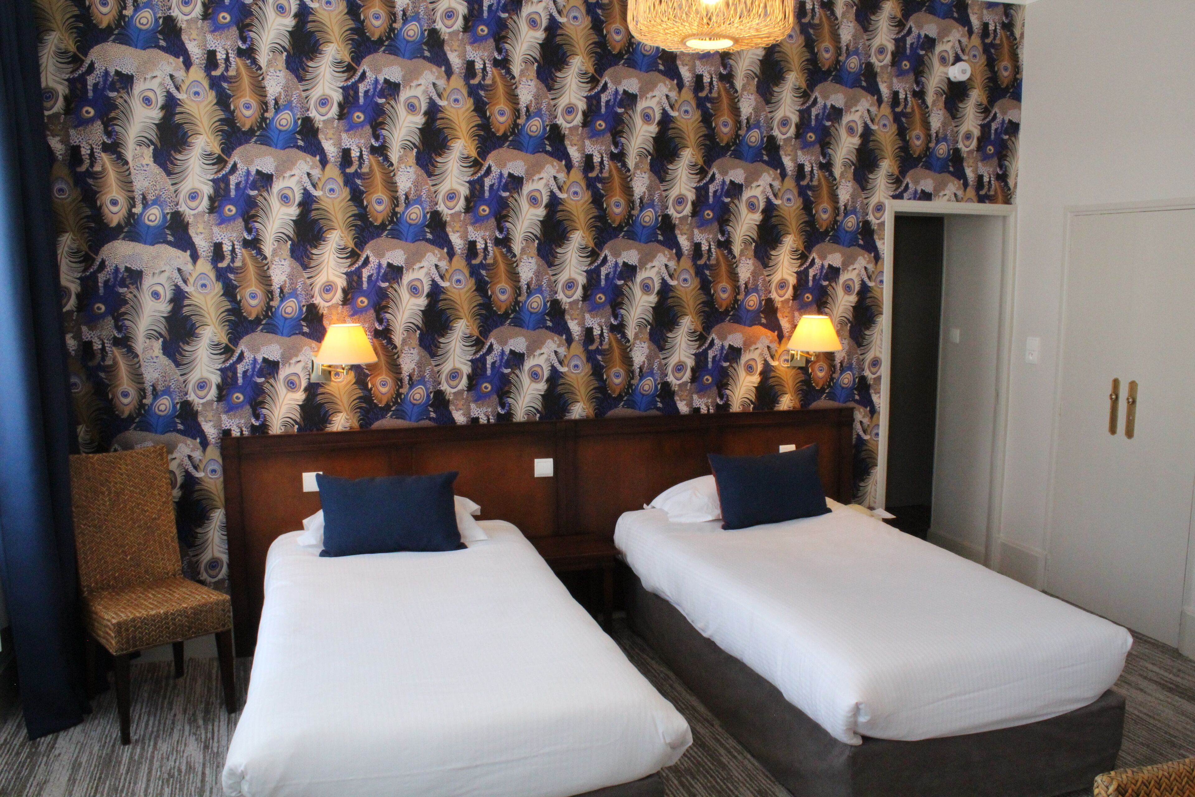 Une chambre d’hôtel avec deux lits simples, une tête de lit en bois, une chaise et un papier peint à motifs.