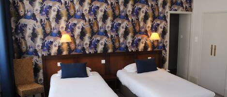 A hotel room with two single beds, a wooden headboard, a chair, and a patterned wallpaper.