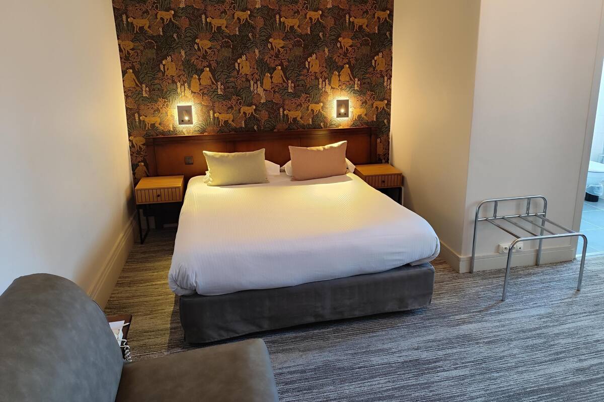 A hotel room with a large bed, two bedside tables, a headboard, a grey sofa, a patterned wall, and a metal rack.