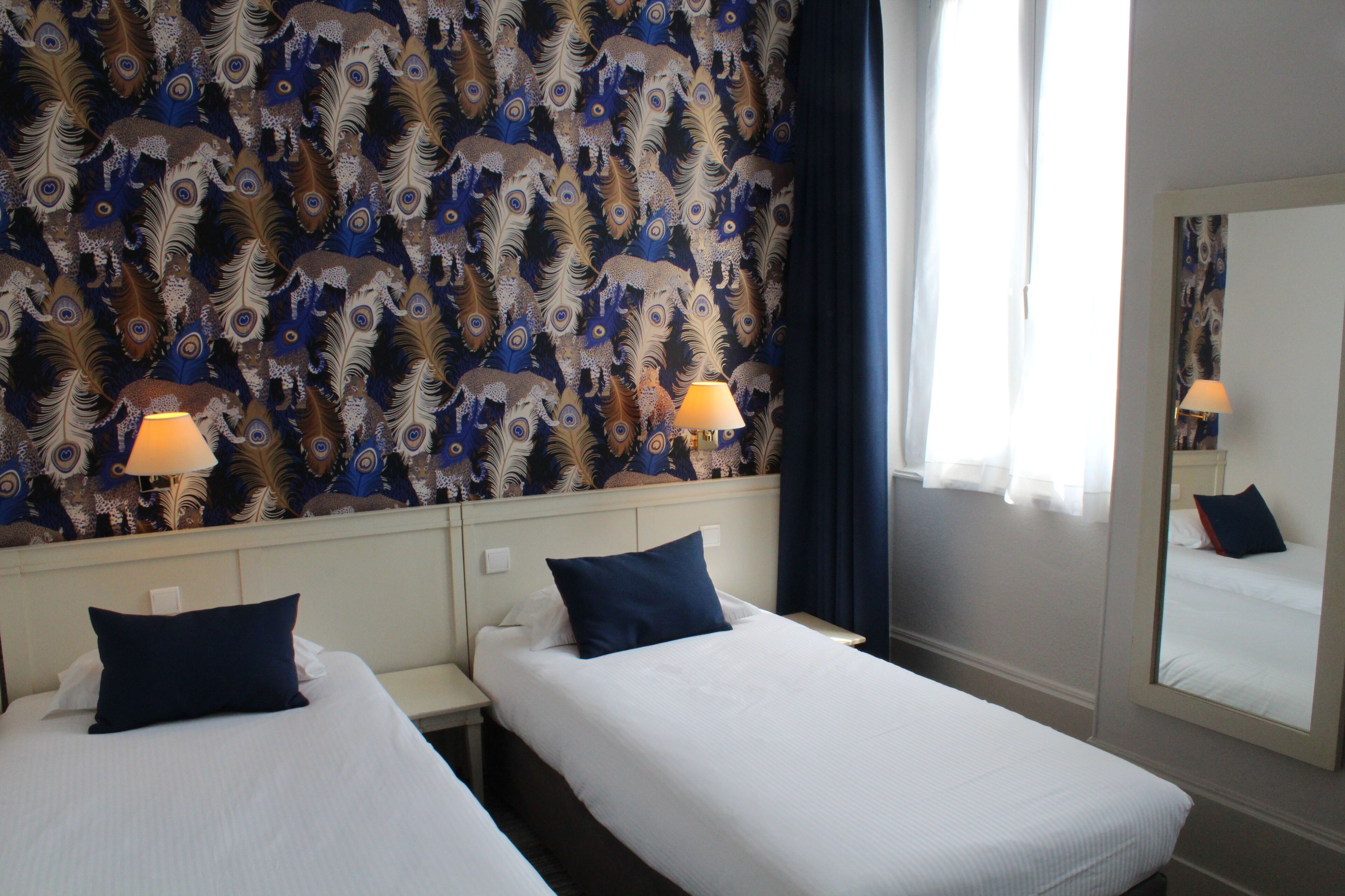 A hotel room with two single beds, a large mirror, and a patterned wallpaper.