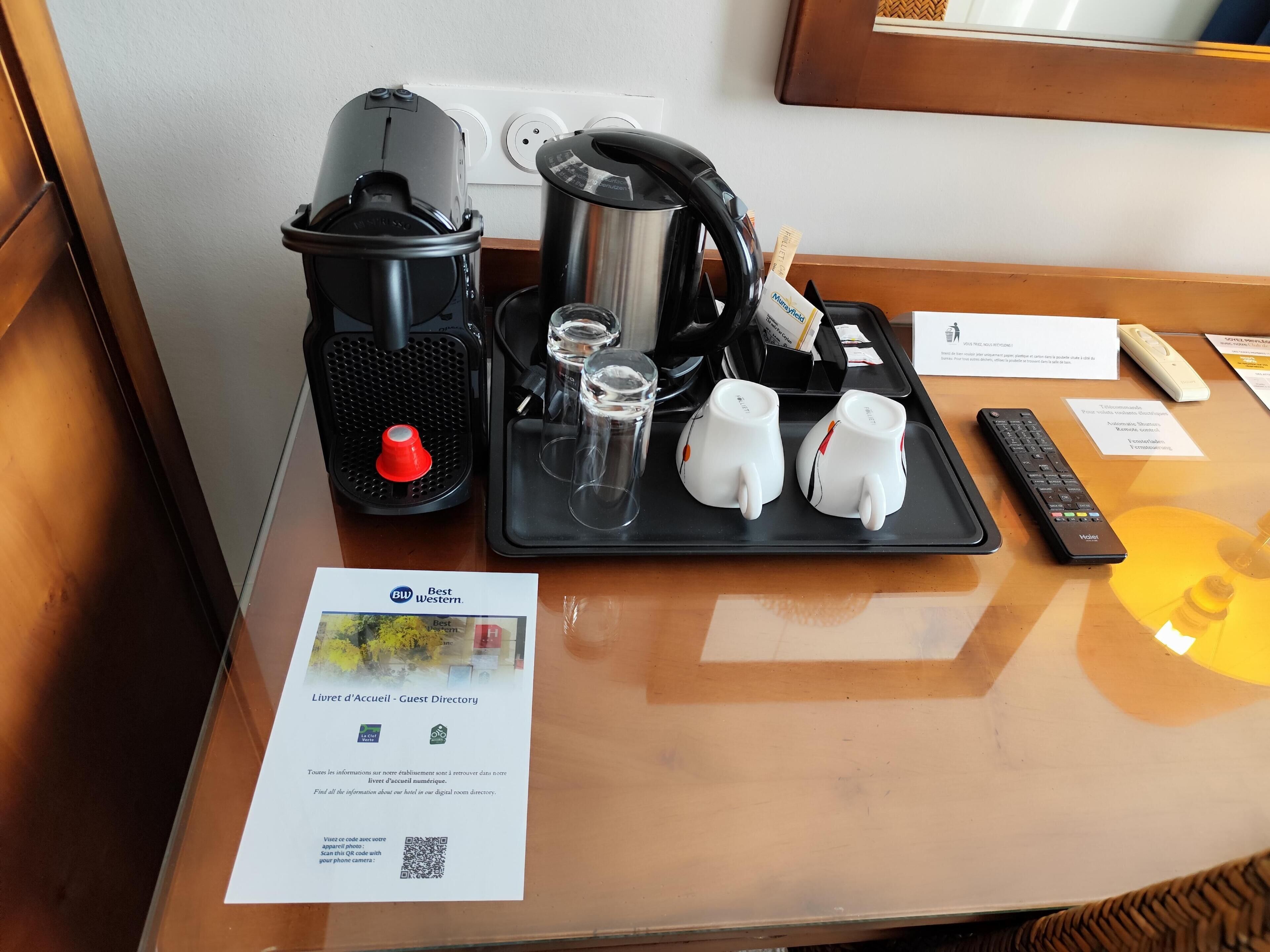 A hotel room's reception area with a coffee maker, tea kettle, glasses, mugs, and a remote control on a tray.