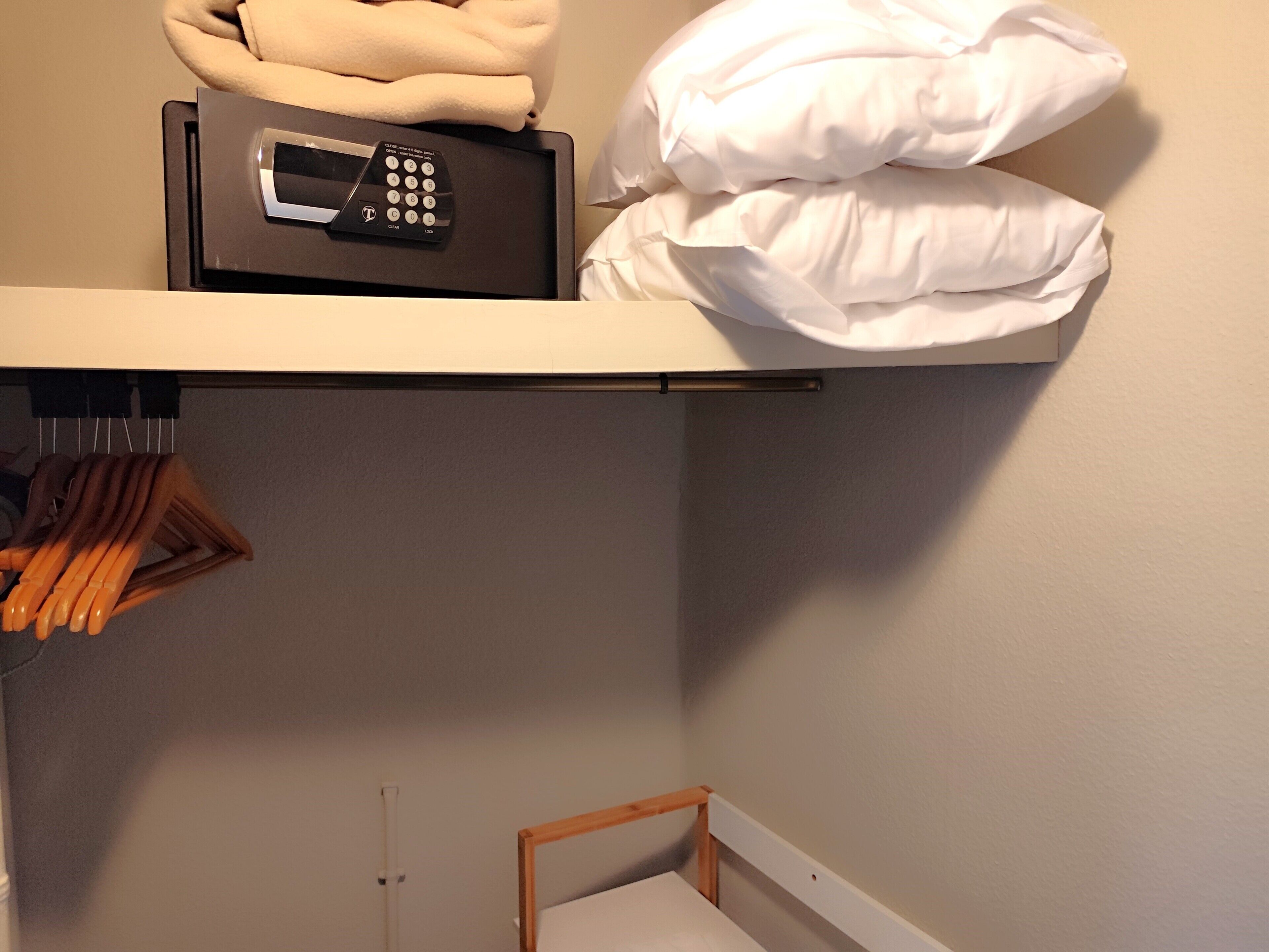 A hotel room with a bed, a bedside table, a wall-mounted shelf with a safe, and a hanging rack with towels.