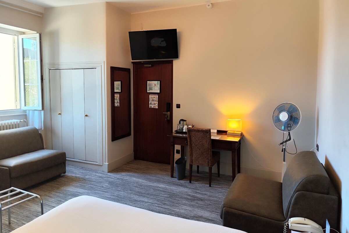 Une chambre d’hôtel comprenant un lit, deux fauteuils, un bureau avec une lampe, une télévision et un ventilateur sur pied.