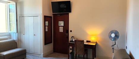 A hotel room with a bed, two armchairs, a desk with a lamp, a television, and a standing fan.