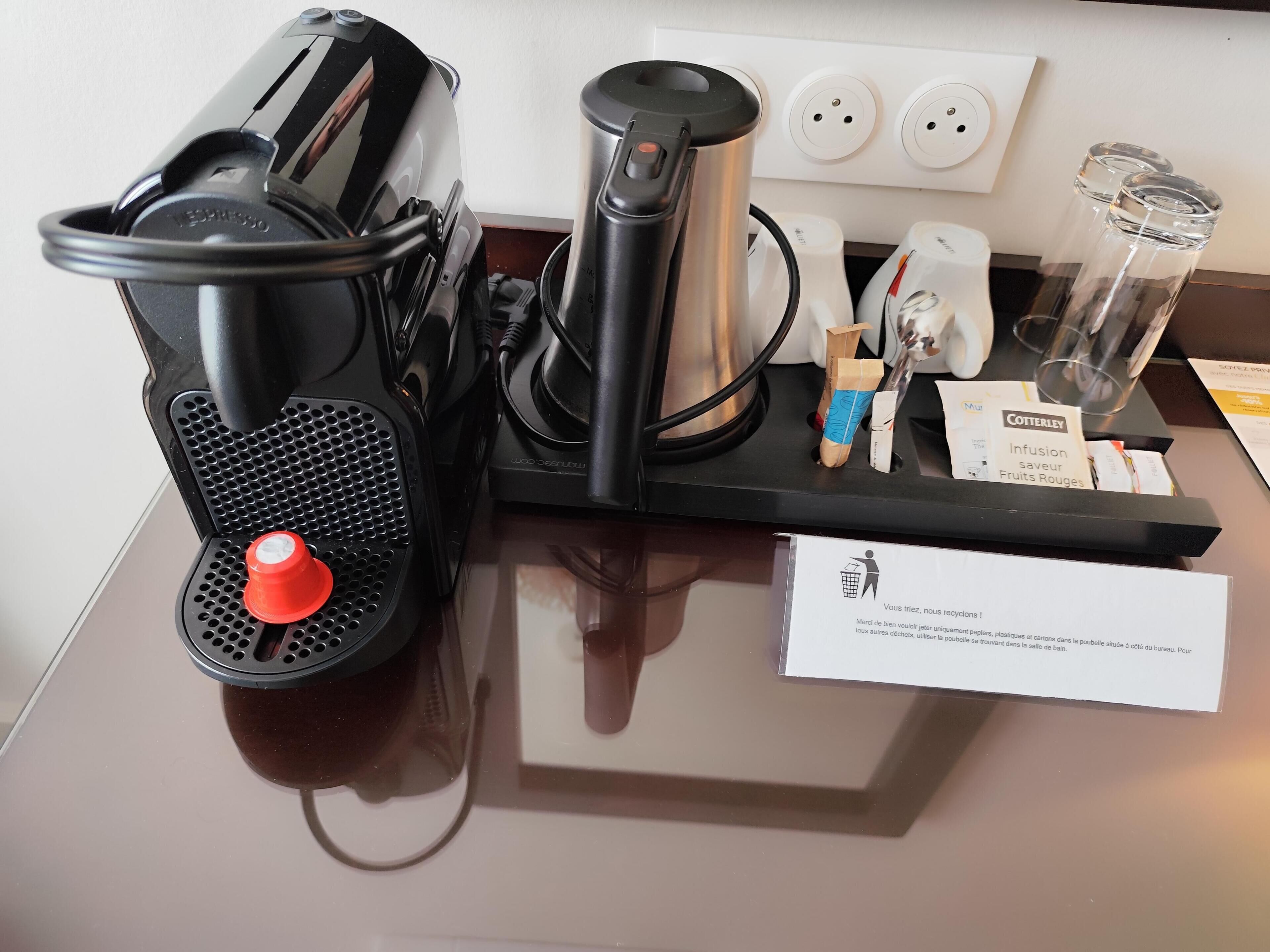 Une machine à café, une bouilloire et un distributeur d’eau sont posés sur un comptoir, à côté d’un panneau indiquant les consignes de tri.