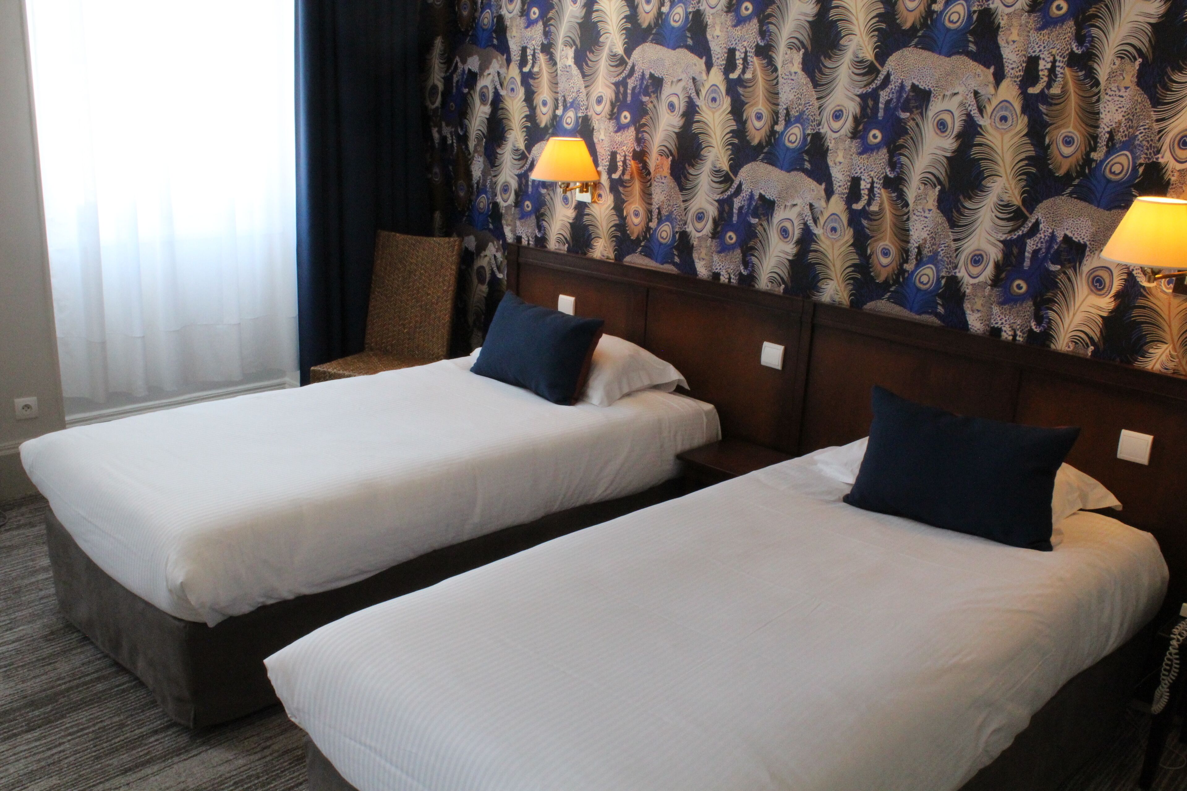 A hotel room with two single beds, a wooden headboard, a patterned curtain, and a lamp on each side.