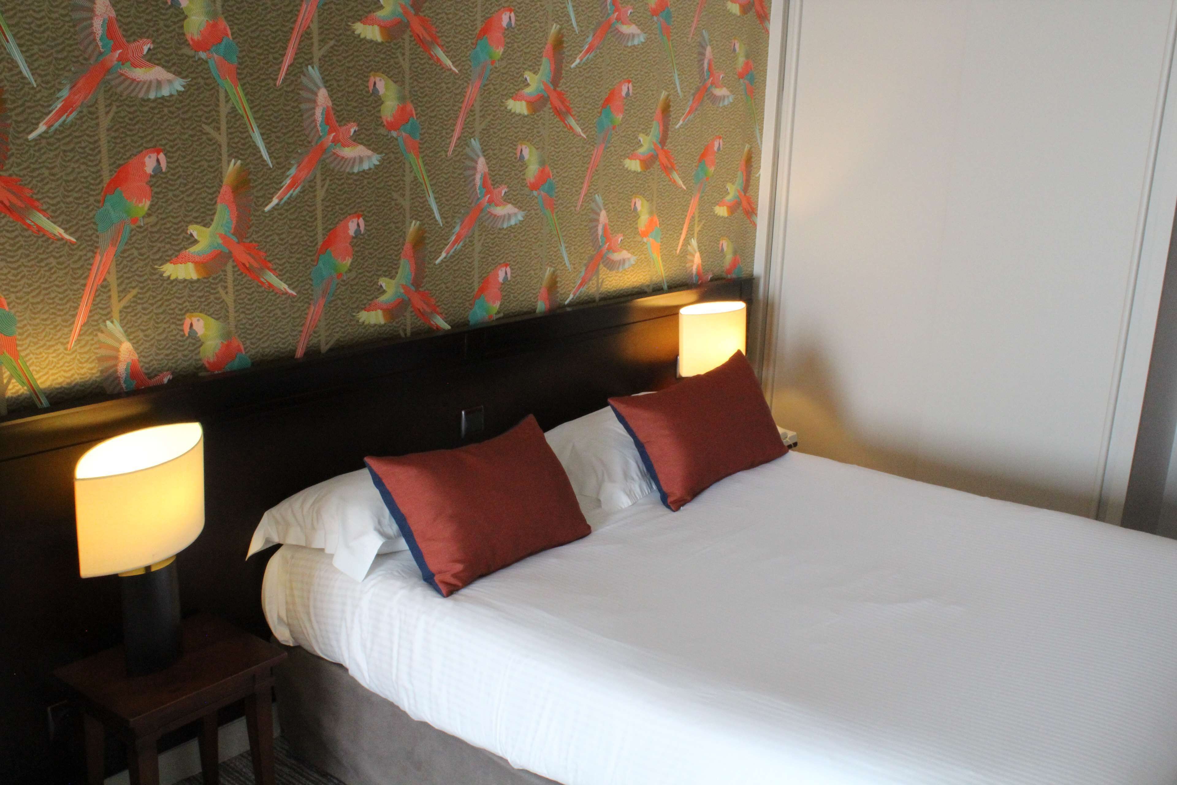 A hotel room with a large bed, two bedside lamps, and a wall with a parrot pattern.