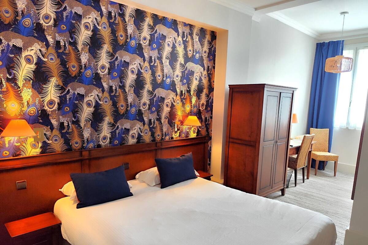 A hotel room with a large peacock feather patterned wall, a bed with white linens, two bedside lamps, a wooden wardrobe, and a dining area with a table and chairs.