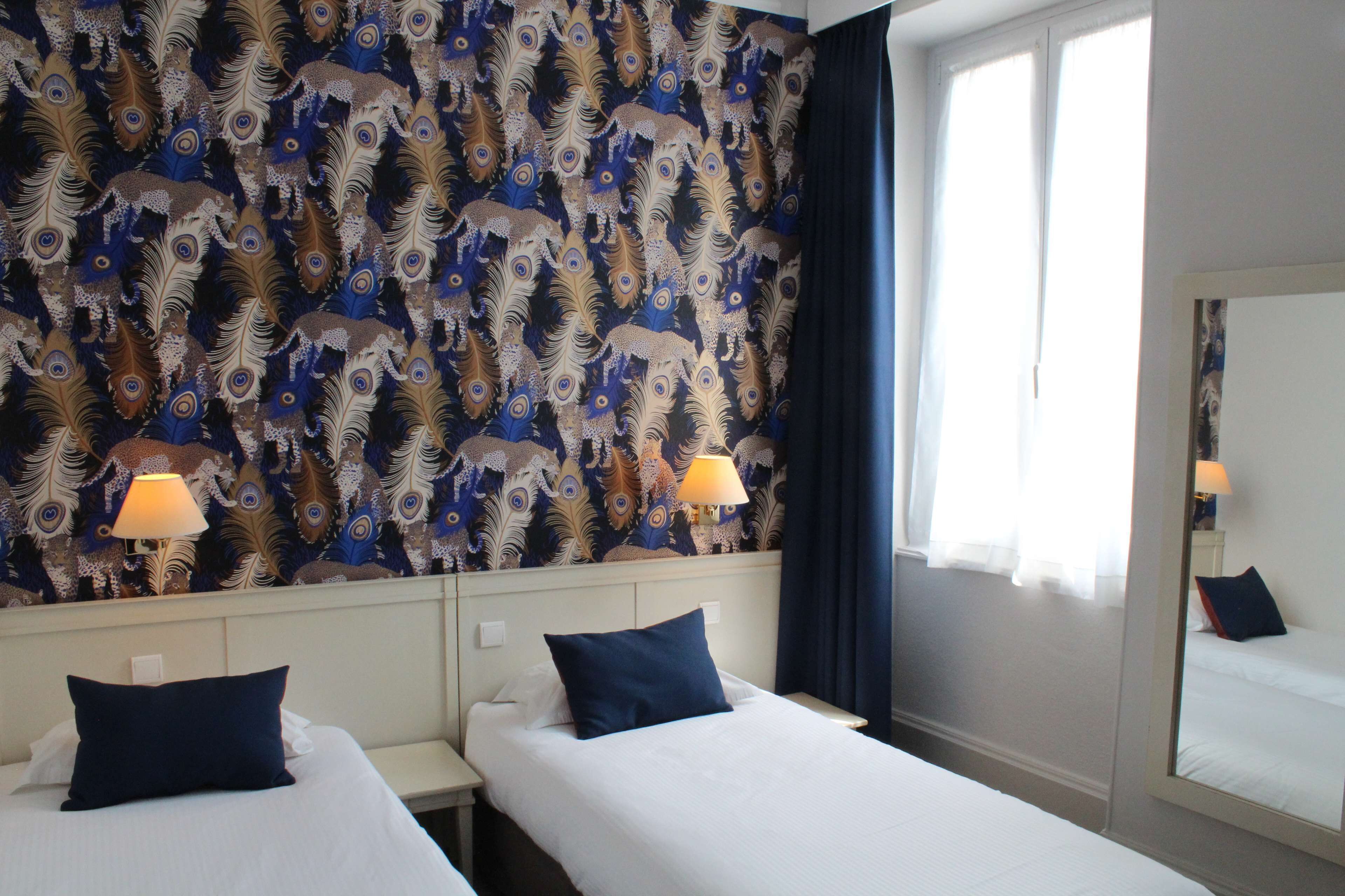 A hotel room with two beds, a patterned wallpaper, a large mirror, and a window with curtains.
