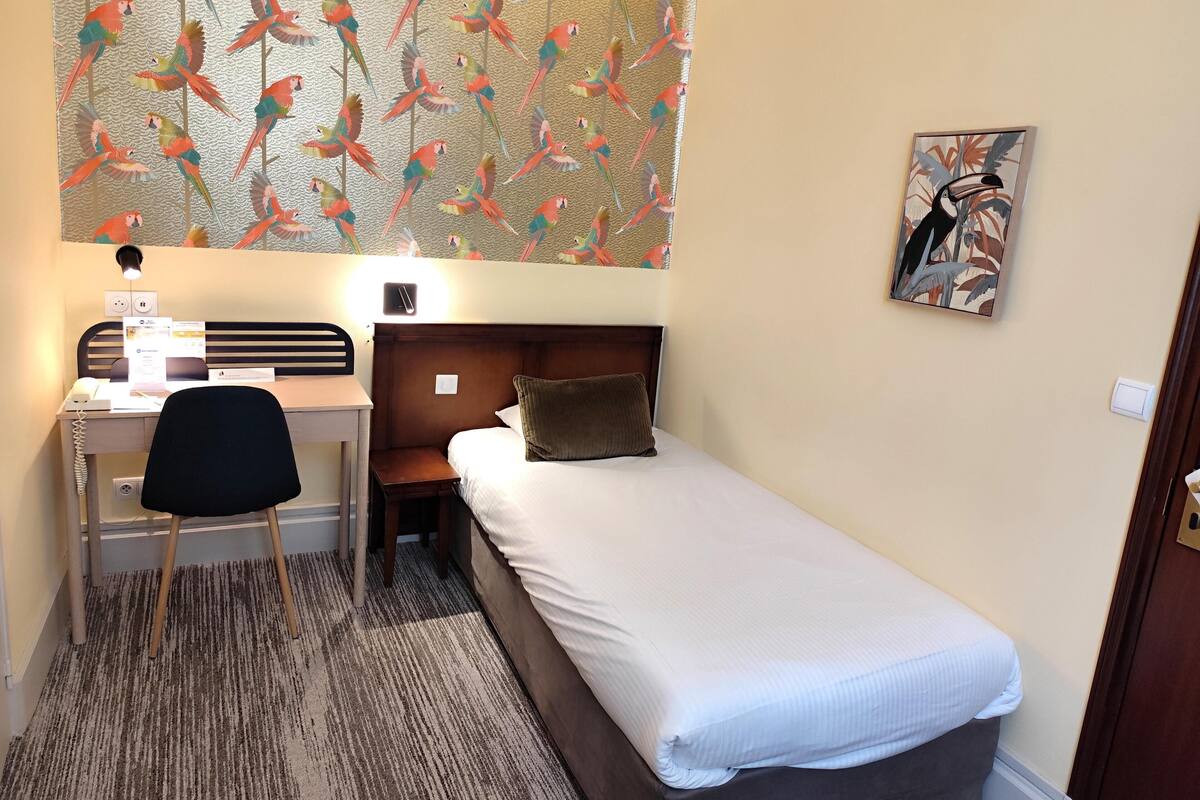 A hotel room with a bed, a desk with a chair, a lamp, a wall with a parrot pattern, and a painting.