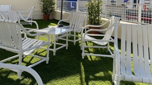 Un patio con sillas blancas, un paraguas grande, una planta pequeña en maceta y un edificio con un cartel que dice "TE".