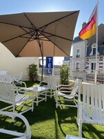 Un patio con sillas blancas, un paraguas grande, una planta pequeña en maceta y un edificio con un cartel que dice "TE".