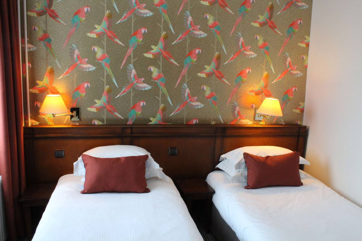 A hotel room with two beds, each with a red pillow and a lamp on either side. The wall behind the beds has a colorful parrot pattern.