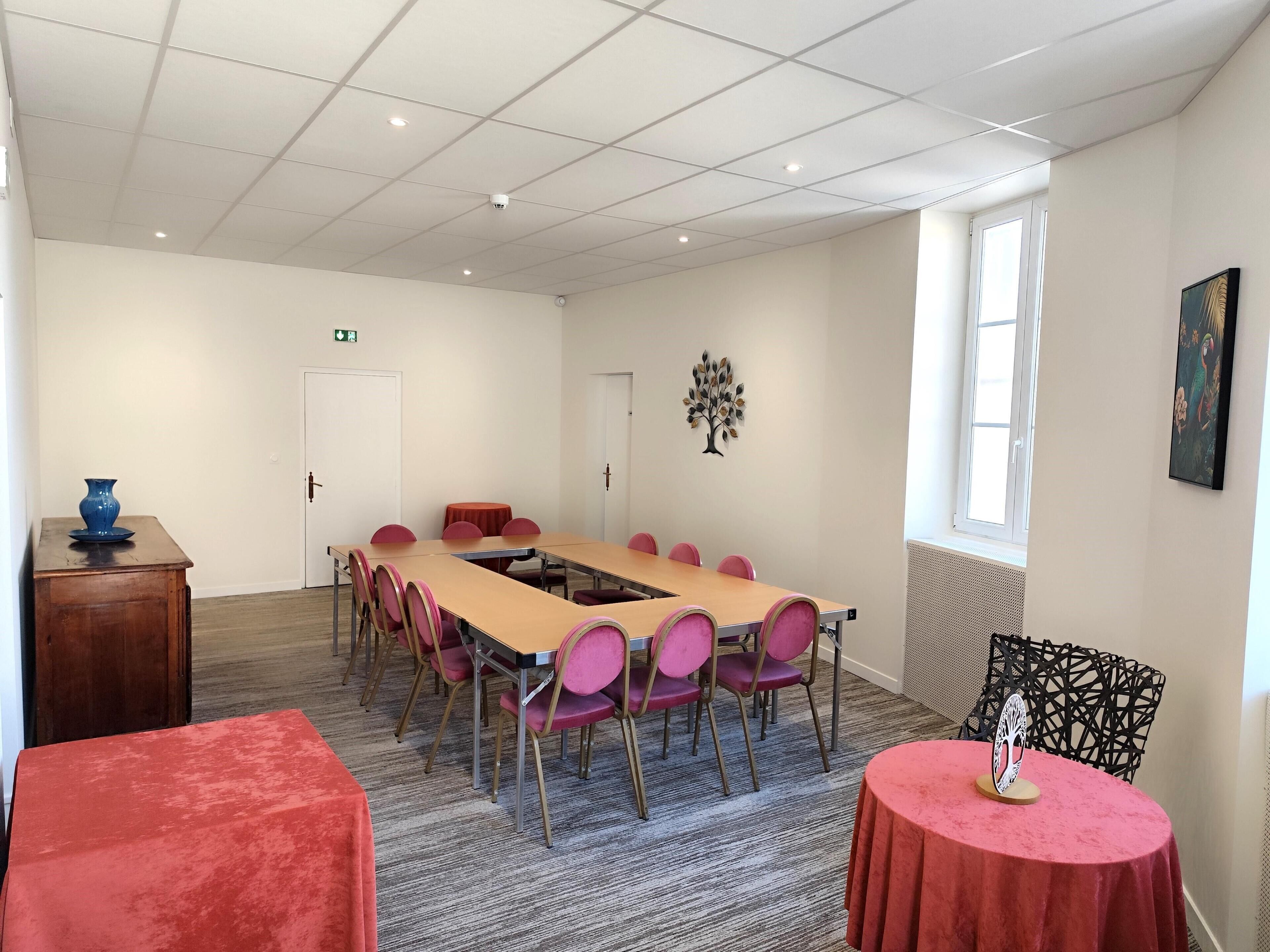 Una sala de conferencias con una larga mesa, sillas, una pequeña mesa con un florero y una puerta.