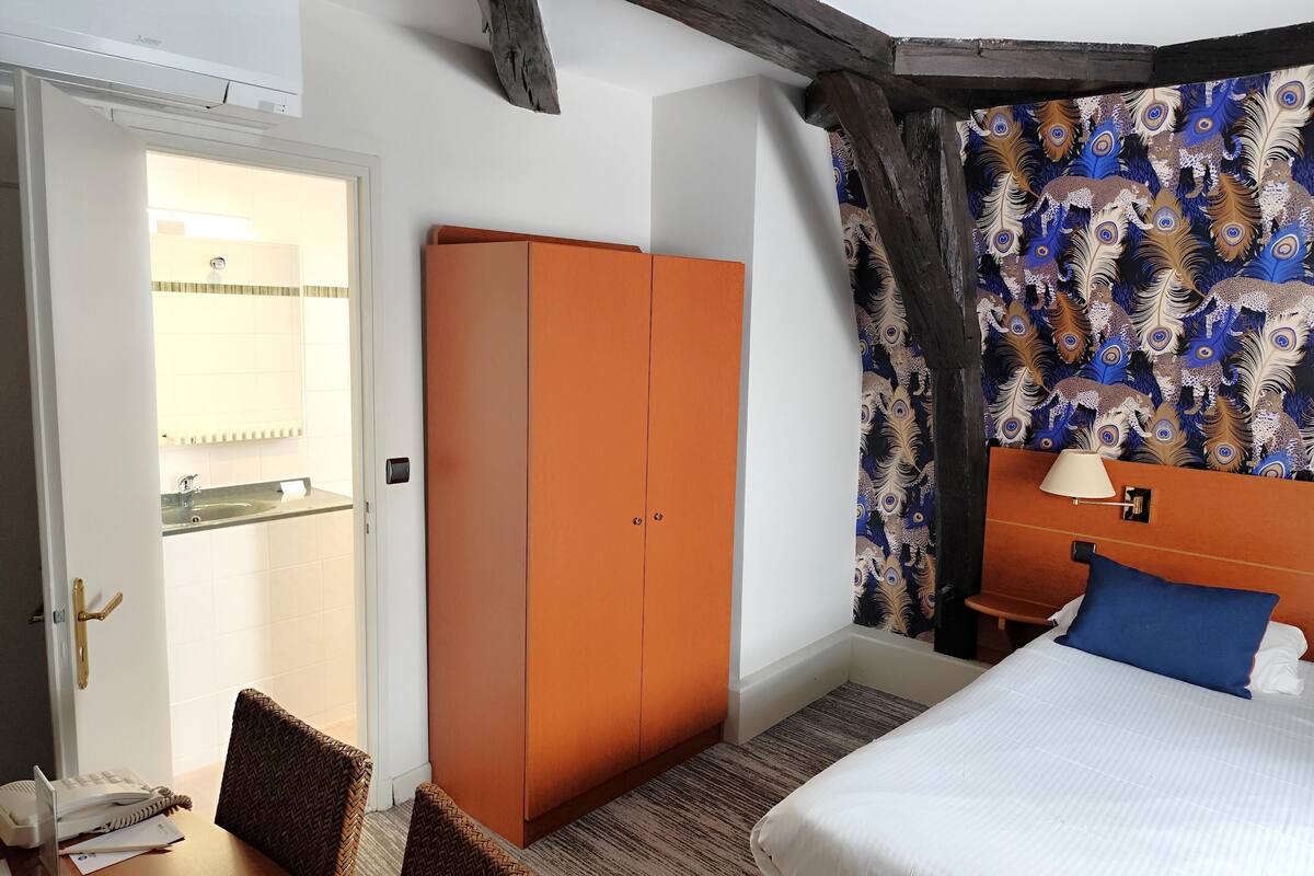 A hotel room with a bed, a wardrobe, a desk with a chair, a bathroom with a sink, and a patterned wall.
