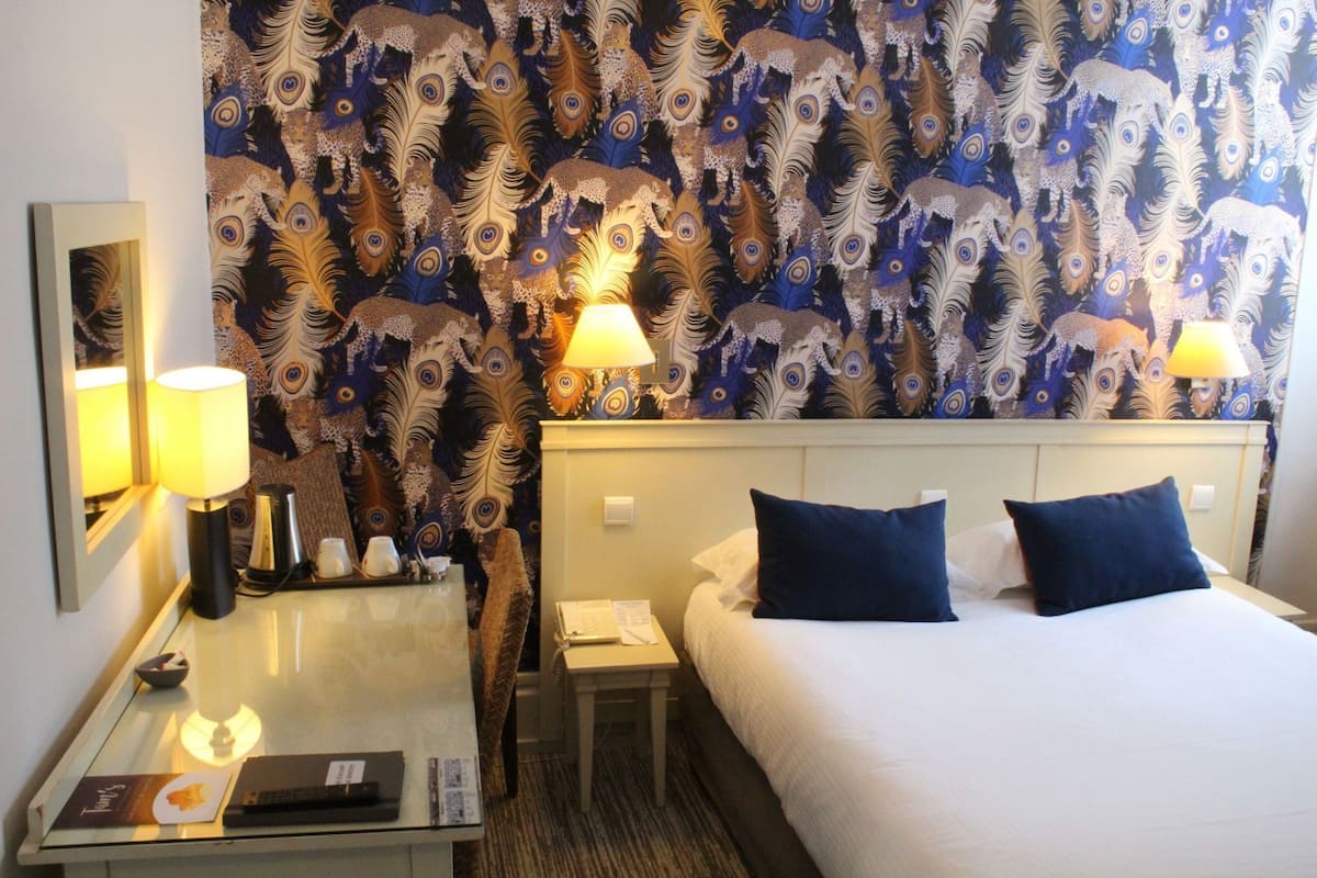 Quarto de hotel com cama, duas luminárias laterais, mesa de cabeceira com telefone e abajur, espelho e mesa com bandeja de objetos.