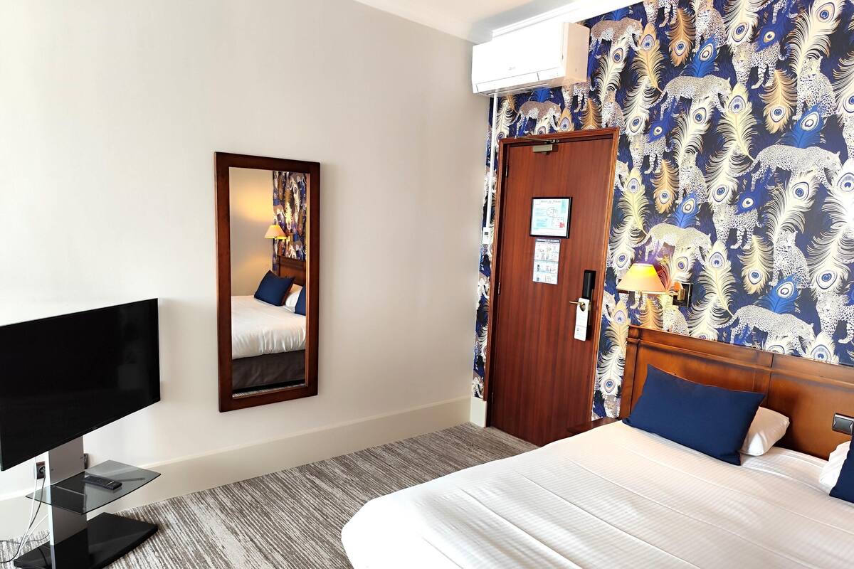 A hotel room with a bed, a TV, a mirror, a door, and a patterned wallpaper.
