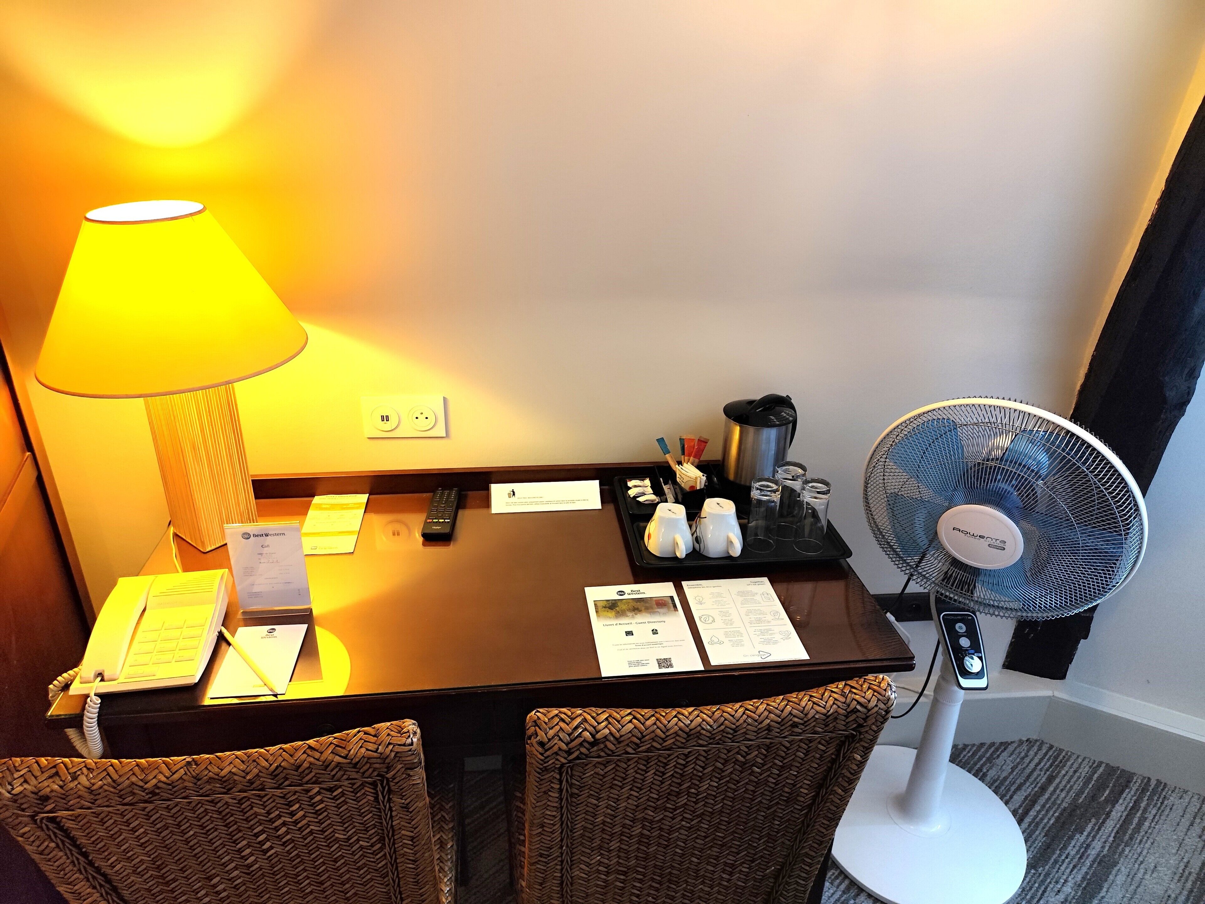 A hotel room with a desk, a lamp, a fan, and a tray with amenities.