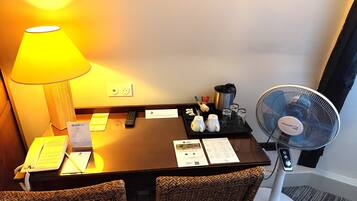 A hotel room with a desk, a lamp, a fan, and a tray with amenities.
