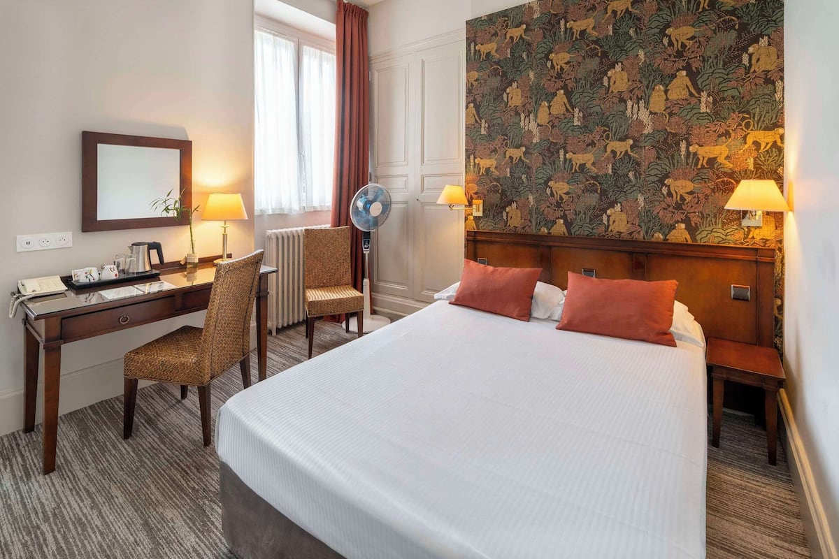 A hotel room with a large bed, a desk with a chair, a standing fan, and a decorative wall.