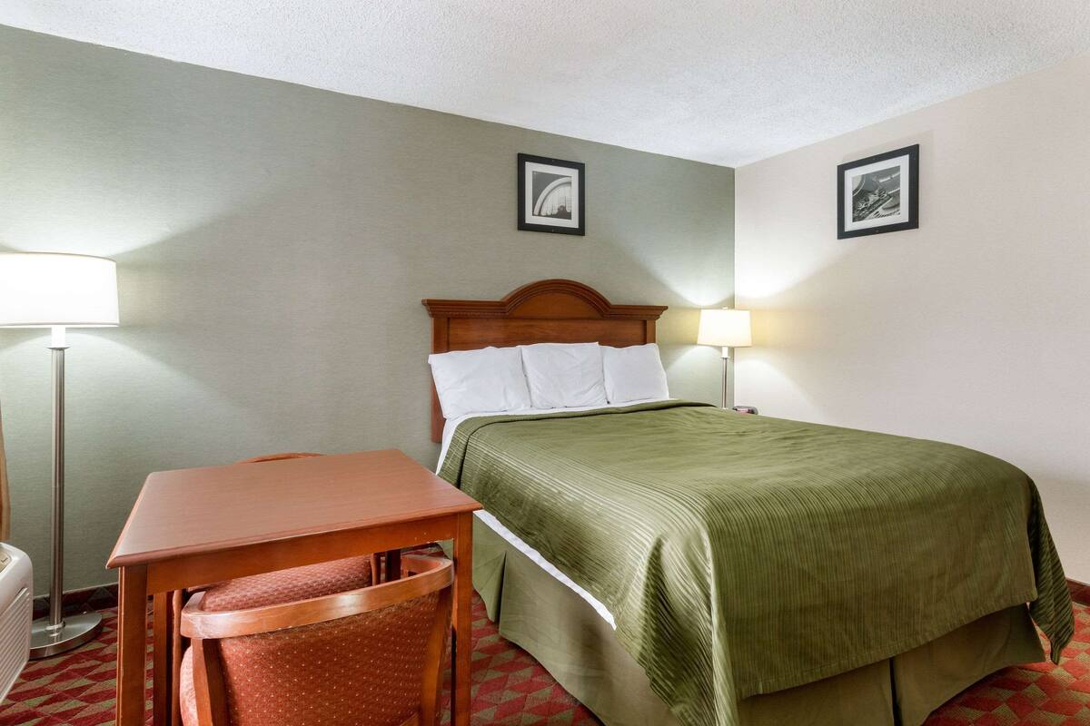 A hotel room with a bed, a wooden nightstand, a desk, and a chair. There are two wall-mounted lamps and two framed pictures on the wall.