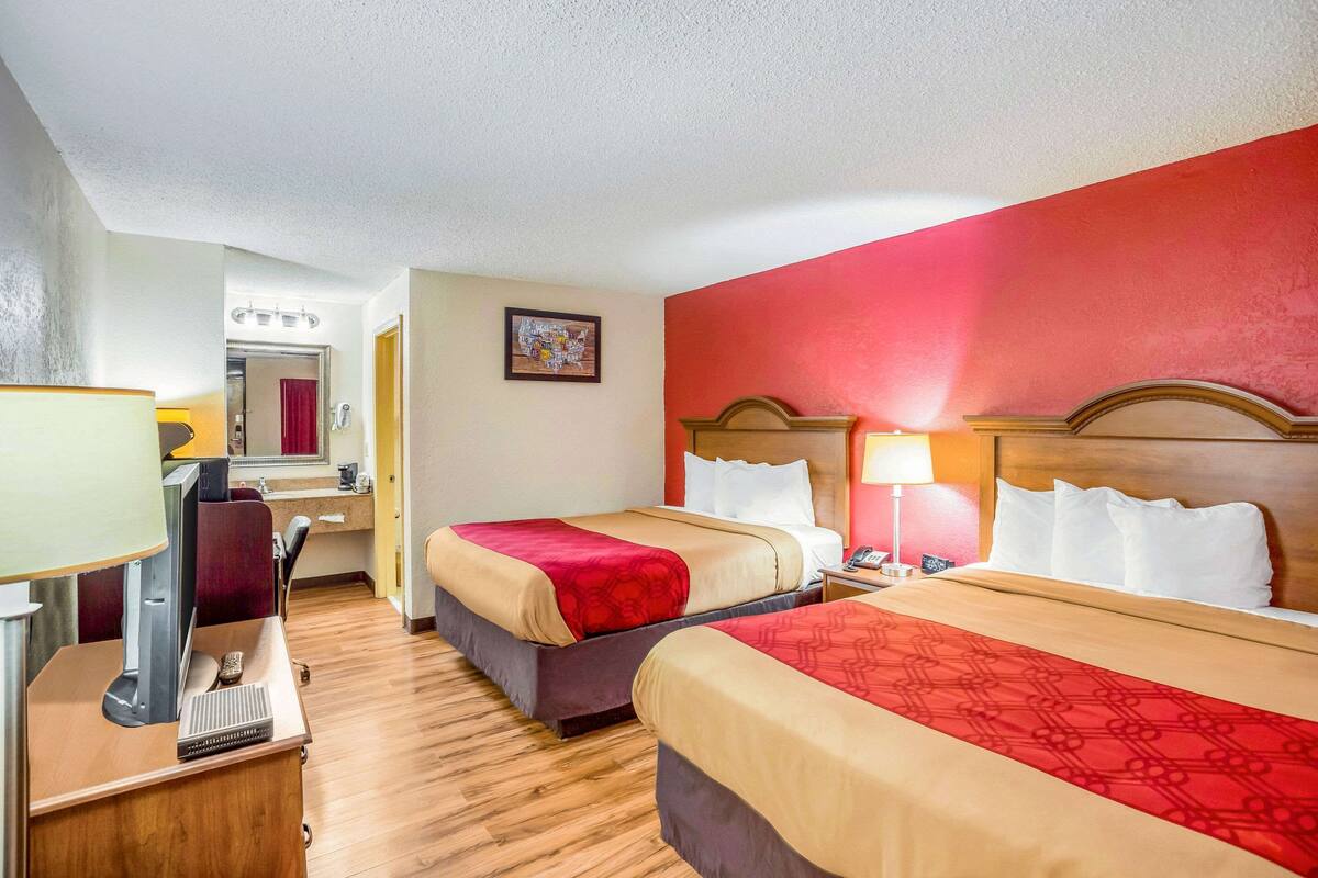 A hotel room with two beds, a desk with a computer, a mirror, and a red wall.