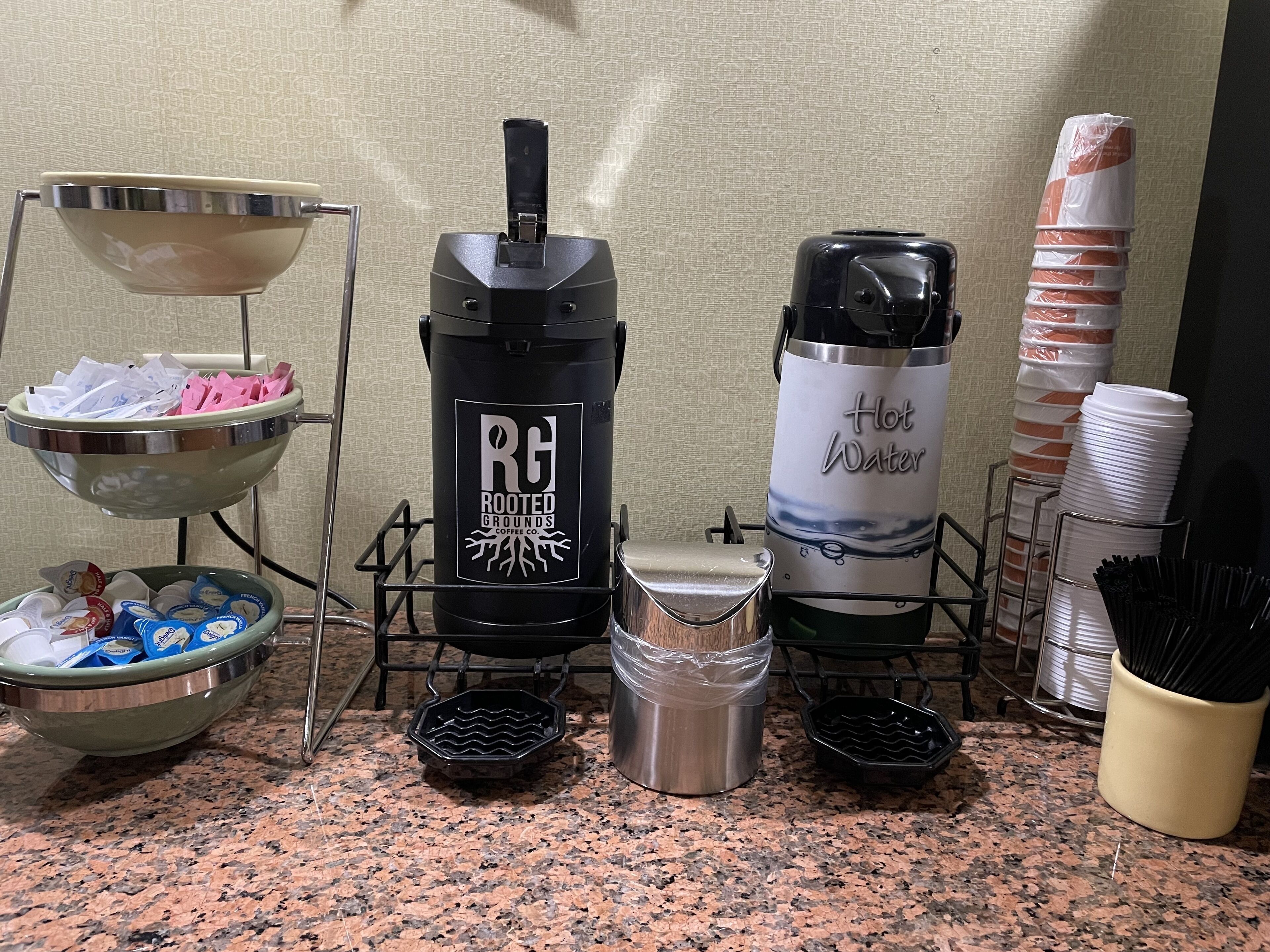 A kitchen area with a coffee maker, a hot water dispenser, a bowl of coffee pods, and a stack of disposable cups.