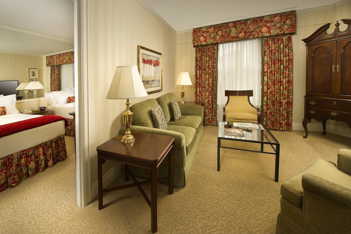A hotel room with a bed, a green sofa, a wooden coffee table, a yellow armchair, a wooden cabinet, and a large window with floral curtains.