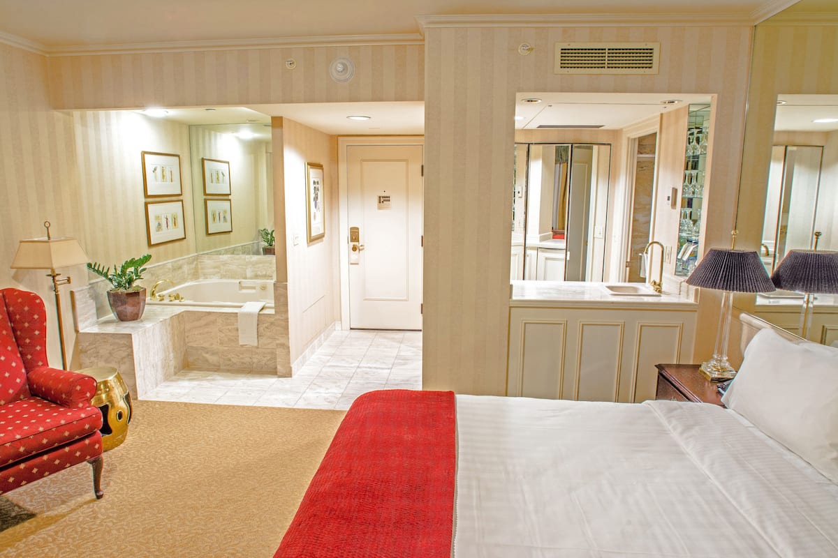 A hotel room with a large bed, a red armchair, a small table with a lamp, a bathroom with a bathtub, and a mirror reflecting the room.