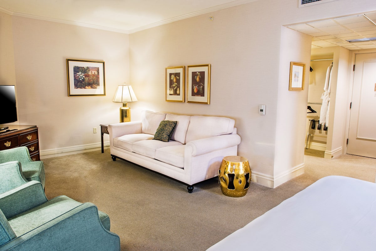 A hotel room with a white sofa, a small side table with a lamp, a gold decorative stool, and a bed.