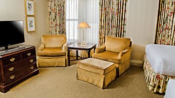 A hotel room with a flat-screen TV, two armchairs, a small table with a lamp, a wooden dresser, and a bed with floral curtains.