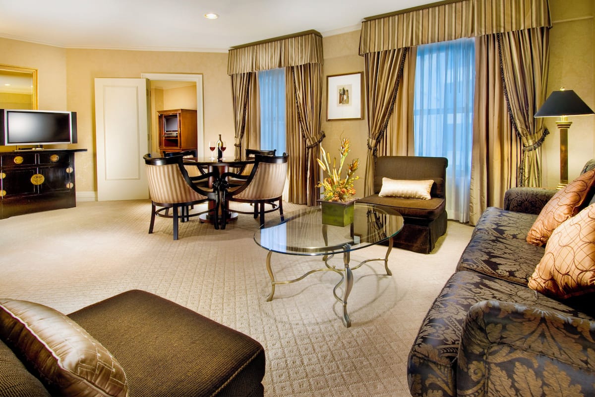 A hotel room with a glass-top coffee table, a sofa, a dining table with chairs, a television, and a lamp.