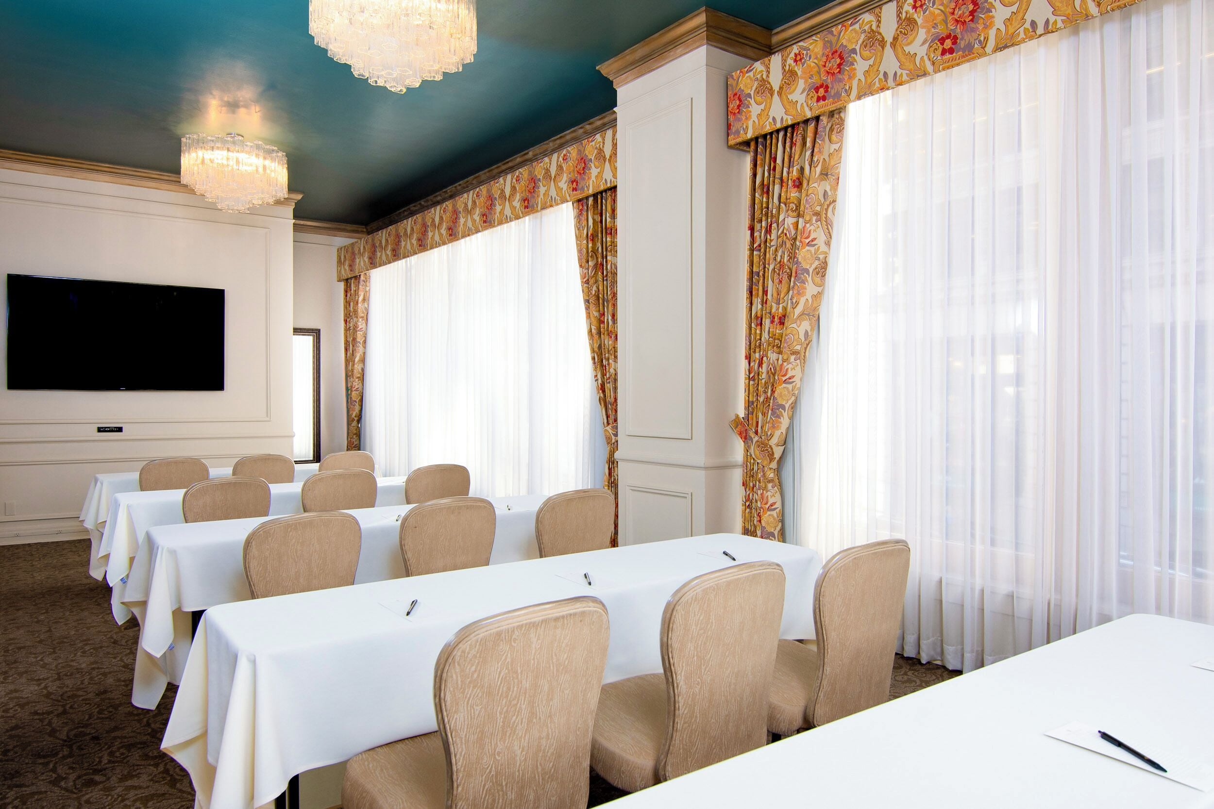 Una sala conferenze con tavoli e sedie bianche, un televisore a schermo piatto e ampie finestre con tende leggere.