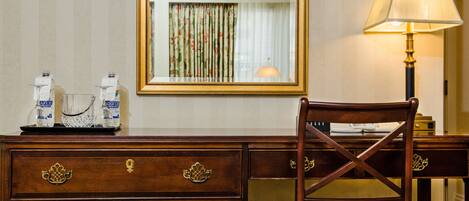 A wooden desk with drawers, a chair with a floral cushion, a lamp, and a mirror reflecting a curtain.