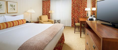 A hotel room with a large bed, a desk with a television, two chairs, and a small table.