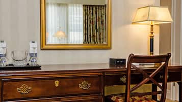 A hotel room with a wooden dresser, a chair with floral upholstery, a lamp, and a mirror reflecting a window with curtains.