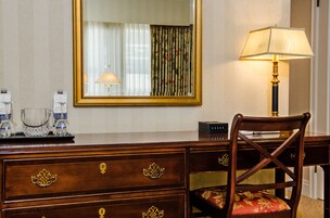 A hotel room with a wooden dresser, a chair with floral upholstery, a lamp, and a mirror reflecting a window with curtains.