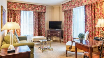 Una habitación de hotel con un sofá verde, un escritorio de madera, un televisor y cortinas con motivos florales.