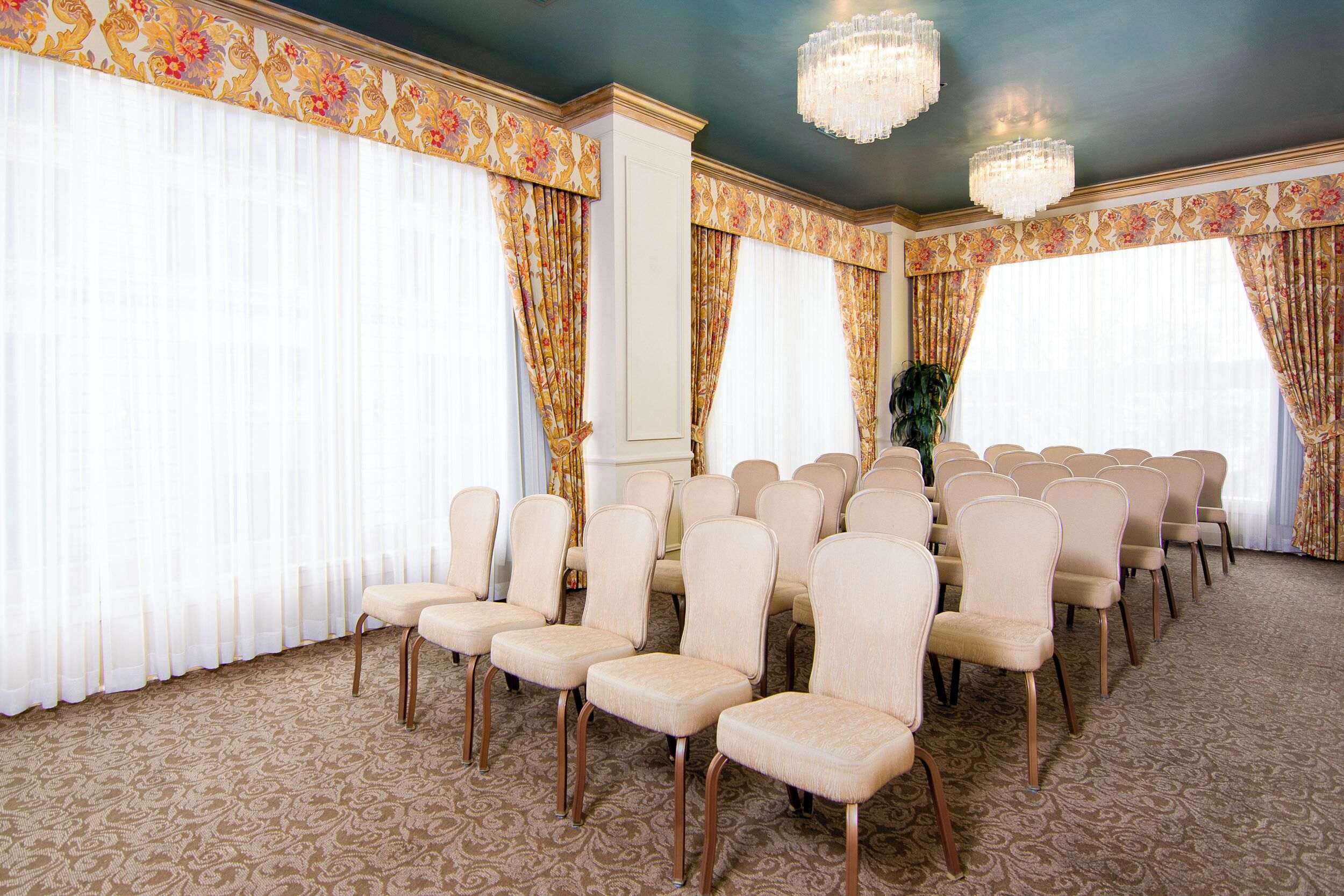 A conference room with rows of beige chairs, large windows with floral curtains, and a patterned carpet.