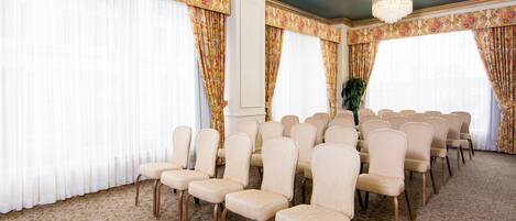 A conference room with rows of beige chairs, large windows with floral curtains, and a patterned carpet.