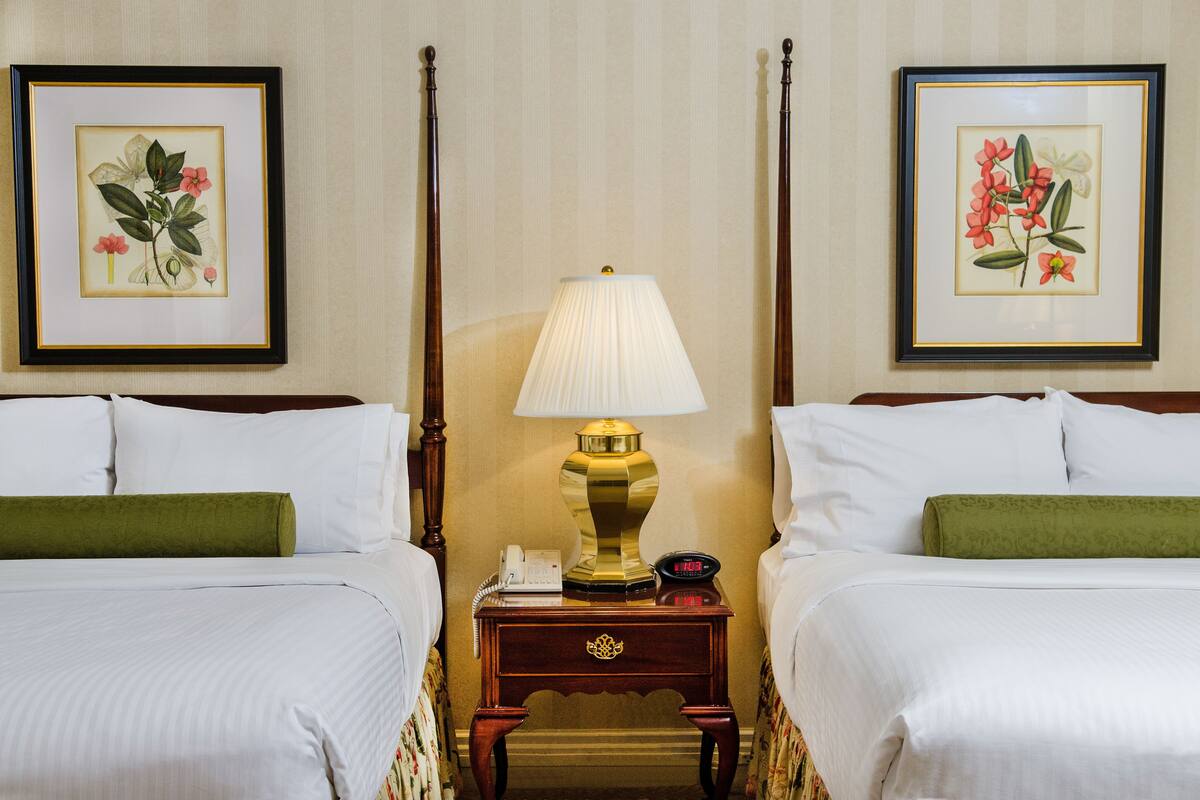 A hotel room with two beds, a nightstand with a lamp, a telephone, and framed botanical prints on the walls.