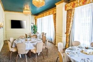 Comedor con mesas redondas dispuestas para una comida, un televisor de pantalla plana en la pared y amplios ventanales con cortinas.