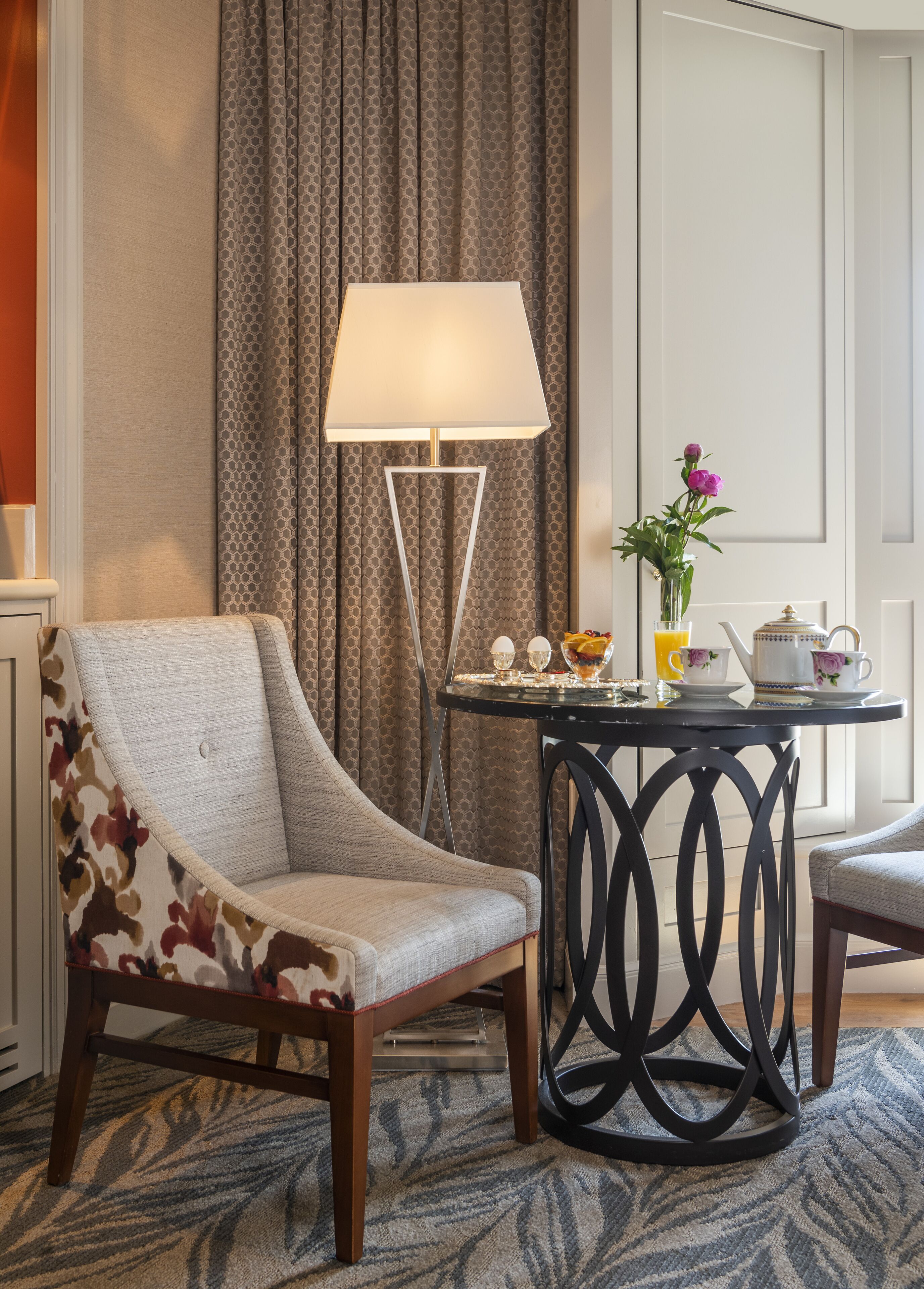 Una camera d'hotel con una poltrona decorata con motivi floreali, un tavolo con un vaso di fiori, una lampada e un servizio da tè.