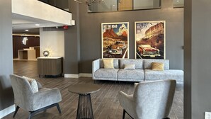 Eine moderne Lobby mit einem grauen Sofa, zwei Sesseln, einem runden Tisch und einer Chandelieber. An der Wand befinden sich zwei Plakate und im Hintergrund ein Empfangsschalter.