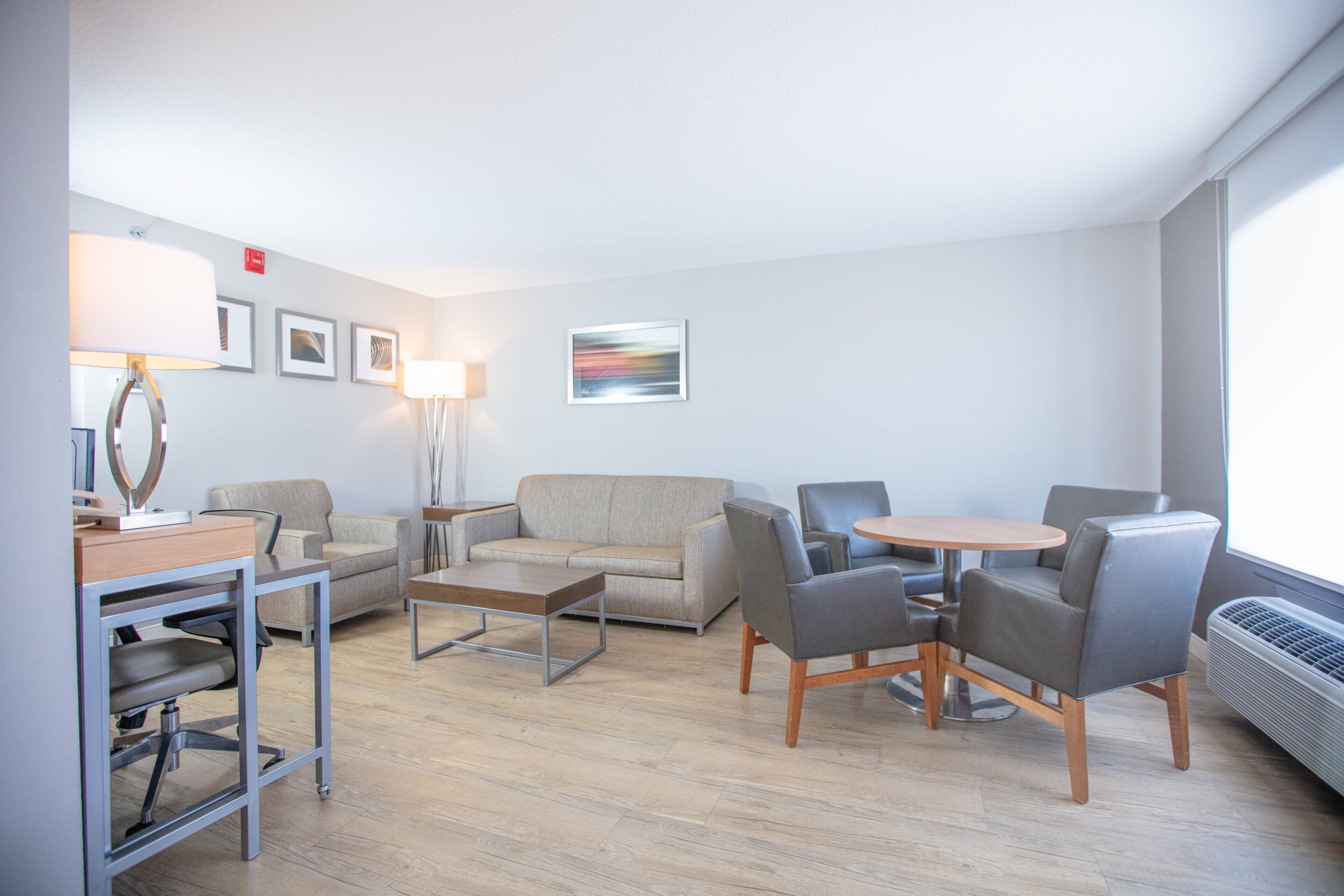 A modern hotel room with a sofa, chairs, a small table, a desk, and a lamp.